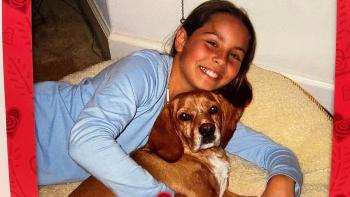 Young girl smiling and holding a brown dog while laying on a floor cushion
