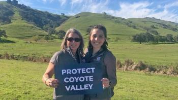two women smiling and holding up a sign that says "protect coyote valley" in front of luscious green hills