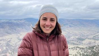 Woman wearing burgundy puffer and grey beanie smiling in front of a scenic view of mountains and green hills