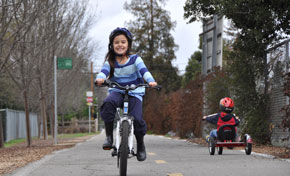 Smiling girl in striped sweater riding a bicycle on paved trail, young boy on tricycle in background 