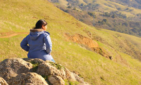 Young woman in blue jacket sitting on rocks taking in view of green hills