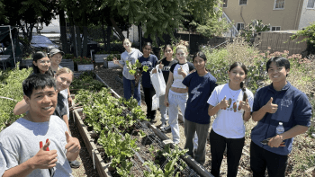 garden box in the center of 2 rows of young people with their thumbs up with trees in the background and a 2 story home on the right side