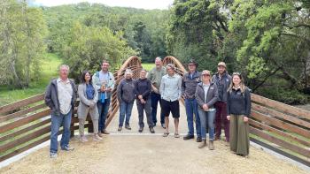 A group of 11 people stand on a dirt trail in front of a pedestrian bridge with leafy tree canopies on either side of the bridge. The people are standing in a horizontal line and smiling at the camera.