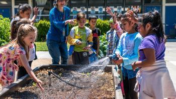 A group of young students in colorful clothing stand smiling and laughing around a raised garden bed, one of the students is holding a sprinkler towards the soil in the bed; a teacher stands to one corner of the bed pointing and smiling at the students