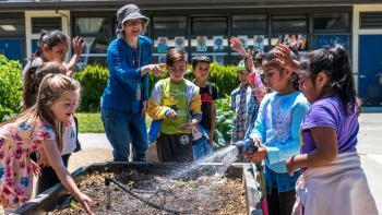 A group of young students in colorful clothing stand smiling and laughing around a raised garden bed, one of the students is holding a sprinkler towards the soil in the bed; a teacher stands to one corner of the bed pointing and smiling at the students