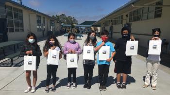 A row of seven children standing outside of a classroom area holding up white paper bags with educational supplies