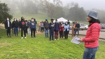 A woman in a red jacket stands in the foreground facing a group of people wearing rain coats standing on a grassy field with a mist-covered hillside in the background
