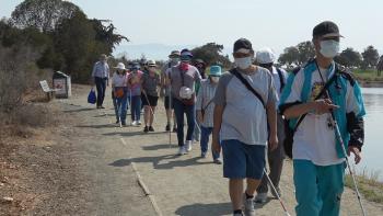 A group of people wearing face masks and most holding mobility canes walking on a paved nature path next to a body of water towards the camera