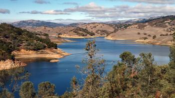 Looking down a covered in green trees towards a blue lake and other golden hills studded with oak trees,, under a blue sky with some white clouds