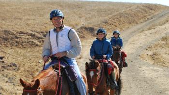 Three horseback riders on a dirt trail through brown hills: one adult in front with two children behind. They are riding towards the camera and smiling, all are on brown horses and wearing helmets.