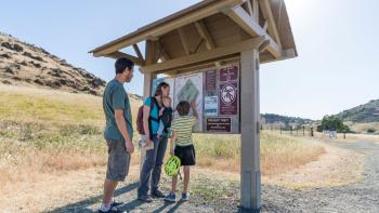 A family of four, two adults, a child, and a baby being carried by the mother, gather around a small trail kiosk with a large map and other informational signage.