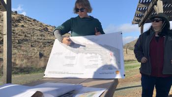A woman in a green shirt holds up and points at a large map of Coyote Valley, with other maps and planning documents on the table in front of her