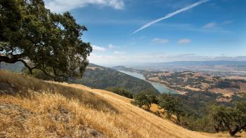 Looking down a hill with golden grass towards a blue lake and other hills studded with oak trees, a valley and mountains far in the distance, under a blue sky with some white clouds