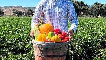 A farmer wearing a white plaid shirt stands in a field full of green leafy crops; he smiles at the camera and holds out a barrel full of orange and red peppers and other vegetables