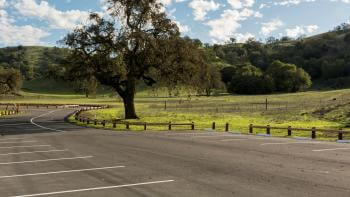 A small, empty parking lot surrounded by a low, split-rail fence next to a green field with a large oak tree and wooded hillsides in the background. The sky is blue and filled with small, fluffy white clouds.