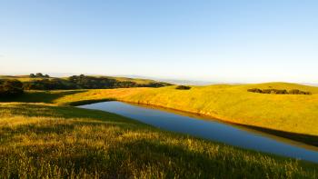 A long blue pond in between golden-green hills under a clear blue sky, with tree-covered hills in the distance