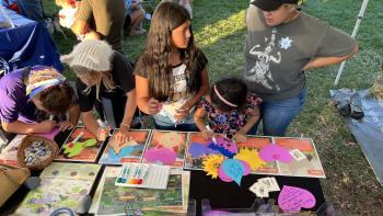 A few children and an adult stand over a table covered in colorful brochures and maps about Coyote Valley, with colorful cut-out shapes that they are writing on