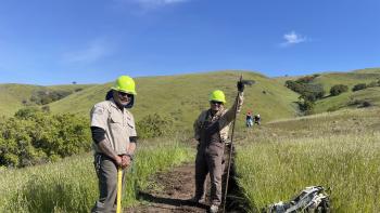 Two field staff members wearing green hard hats and holding trail tools stand smiling at the camera on a dirt trail they are building, in the middle of green grassy hills