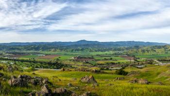 Looking over green hills across a valley below filled with agricultural fields, to blue mountains in the distance, under a blue sky streaked with white clouds