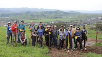 19 volunteers holding yellow road rakes stand smiling at the camera on green grass and a muddy trail, below them is a lush green valley with mountains in the distance