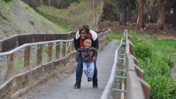 A mother bends over and holds up her small child, both are smiling at the camera. They are standing on a paved trail with wooden railings surrounded by trees and grass.