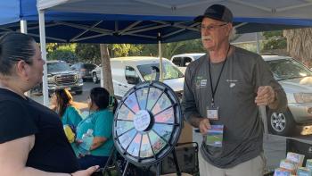A volunteer wearing a dark gray Open Space Authority shirt stands under a booth behind a table full of colorful papers and brochures, he is talking to a visitor standing on the other side of the table