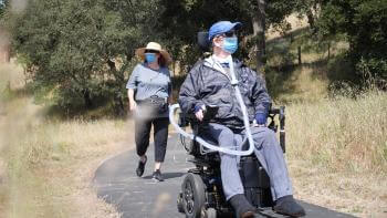 A person wearing a blue face mask in a motorized wheelchair is followed by a person also wearing a blue face mask walking on a paved nature trail. 