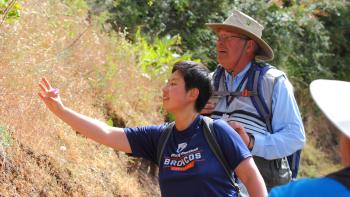 A person wearing a navy blue shirt reaches out to touch a plant on the side of the trail, behind them is an OSA volunteer docent wearing a light blue shirt and tan hat.