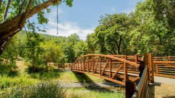 A rust-colored pedestrian bridge spans a small creek surrounded by lush trees on either side and under a bright blue sky