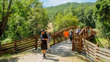 A group of people holding white mobility canes walk across a pedestrian bridge and onto a trail. In the background are tree-covered hills and a blue sky.
