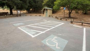 Looking down on a parking lot with two accessible parking spots and a "No Parking" space between them; in the background is a small restroom, a split-rail fence, trees, and a trail kiosk.