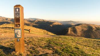A trail marker in the font left side of the photo points towards a trail leading down a grassy hillside towards rolling green mountains in the distance, under a clear light blue sky