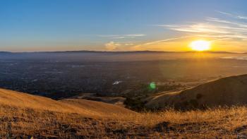 Looking down a hillside covered in golden grass across the South Bay Area to dark hills where a bright yellow sun is setting over a blue sky