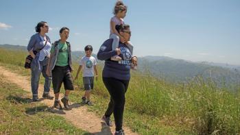 A family with two adults and 3 children walks towards the camera on a dirt trail surrounded by green grass with mountains in the background, under a blue sky. The adult in the front is carrying one of the children on her shoulders.