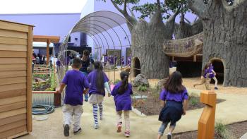 Children wearing dark purple t-shirts running towards an outdoor play area with tree-shaped play structures, a rope bridge, and garden planters