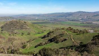 Rolling green hills with oak trees and winding trails overlooking a wide green valley beyond them, and mountains on the far side, under a blue sky.