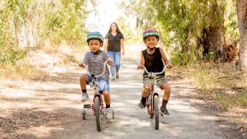 Two smiling young children wearing Teenage Mutant Ninja Turtle helmets ride on small tricycles on a dirt path towards the camera, their mother is out of focus walking behind them