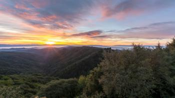 Looking across dark, tree-covered mountains towards a sun rising over misty mountains on the horizon. The sky is light blue with pink and yellow clouds.