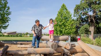 A child wearing pink shorts, a white shirt, and purple ear protection headphones walks across a play structure make up of logs, holding the hand of her father who is wearing jeans and a black and gray shirt. Behind them is a grassy park and blue sky.