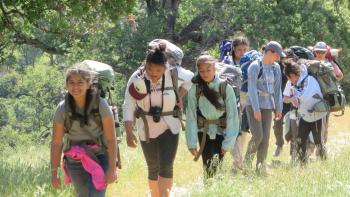 A line of girls wearing backpacking backpacks walk along a trail bordered by tall grass, smiling at the camera