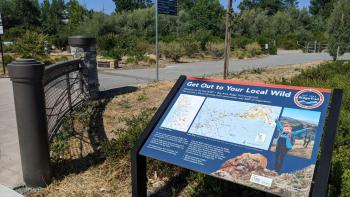 A large educational panel with the title "Get out to your local wild" above a map of the Bay Area, next to a paved trail and wayfinding signage