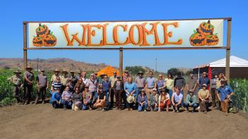 A group of about 40 people standing in a pumpkin patch and smiling at the camera under a large banner that says "WELCOME"