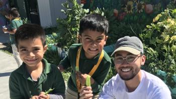 Two young children in green polo shirts stand next to a kneeling young adult in a white t-shirt, holding up pea pods and smiling