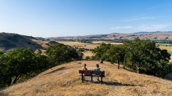 Two hikers sit on a bench overlooking golden hills and trees