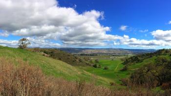 A panoramic photo of green hillsides with hiking trails overlooking a wide green valley, with mountains in the distance, under a blue sky with white fluffy clouds