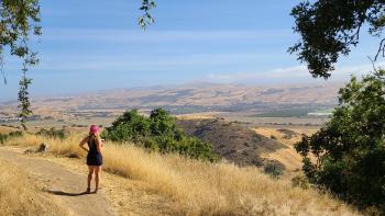 A hiker wearing shorts, tank top, and pink baseball hat stands on a dirt trail looking at a view of a golden valley and open sky