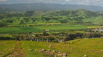 A line of hikers walking along a green ridge top with grass and rocks in front of a beautiful view of a green valley with green hills on the horizon