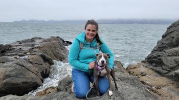 Elizabeth, wearing a turquoise sweatshirt, knees next to a white and gray dog on a rocky outcropping next to the ocean, smiling at the camera