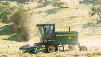 A person on a tractor drives through a hay field with golden hills in the background