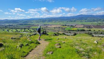 Hiker on a trail on a green grassy field overlooking a beautiful view of a valley patchworked with farm fields and blue mountains in the distance